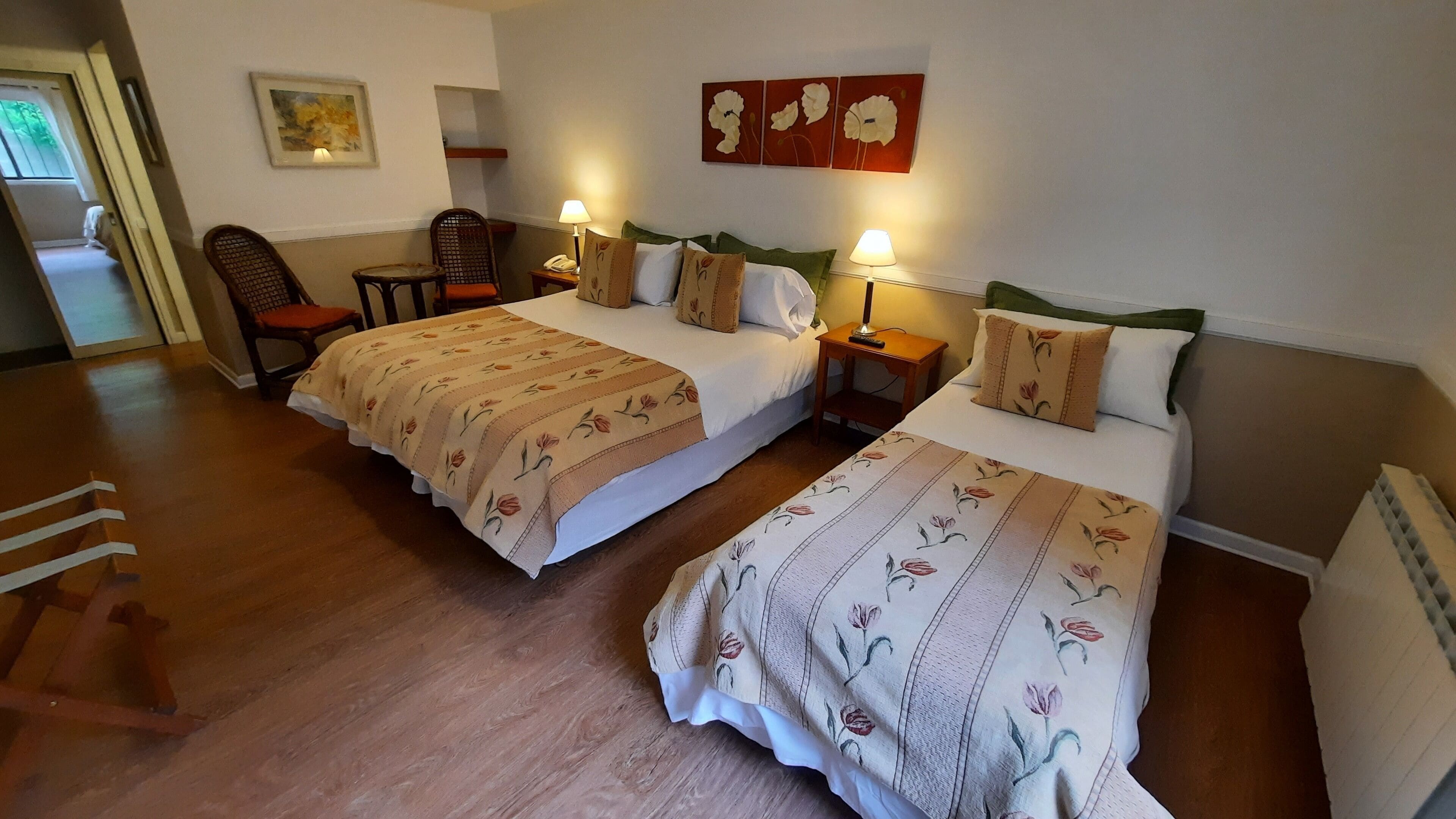 Habitación de hotel con dos camas, suelo de madera, una silla, mesitas de noche con lámparas y cuadros con motivos florales en las paredes.