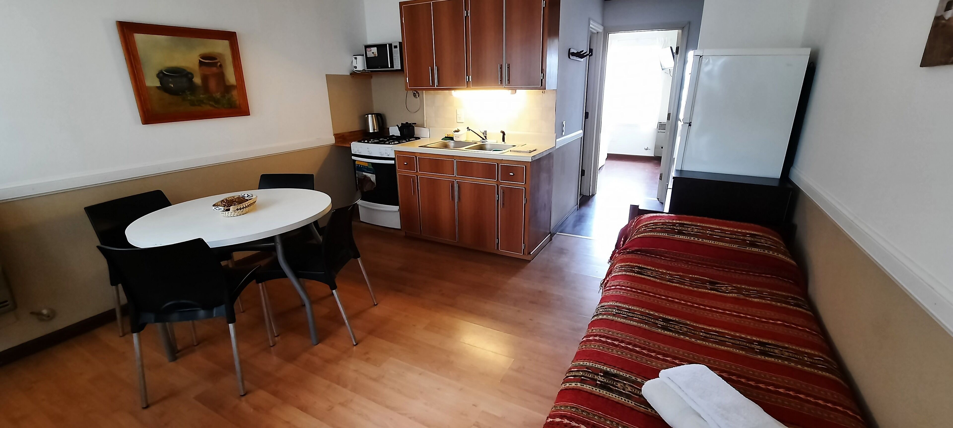 A compact room with a dining table, chairs, a sofa, a kitchen area with cabinets and appliances, and a painting on the wall.