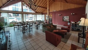 Un espacio interior amplio con vigas de madera, suelo de baldosas y varios conjuntos de asientos, incluyendo mesas y sofás.