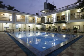 Una piscina con sillas de descanso y sombrillas, rodeada por un edificio con múltiples balcones y suelo a cuadros.