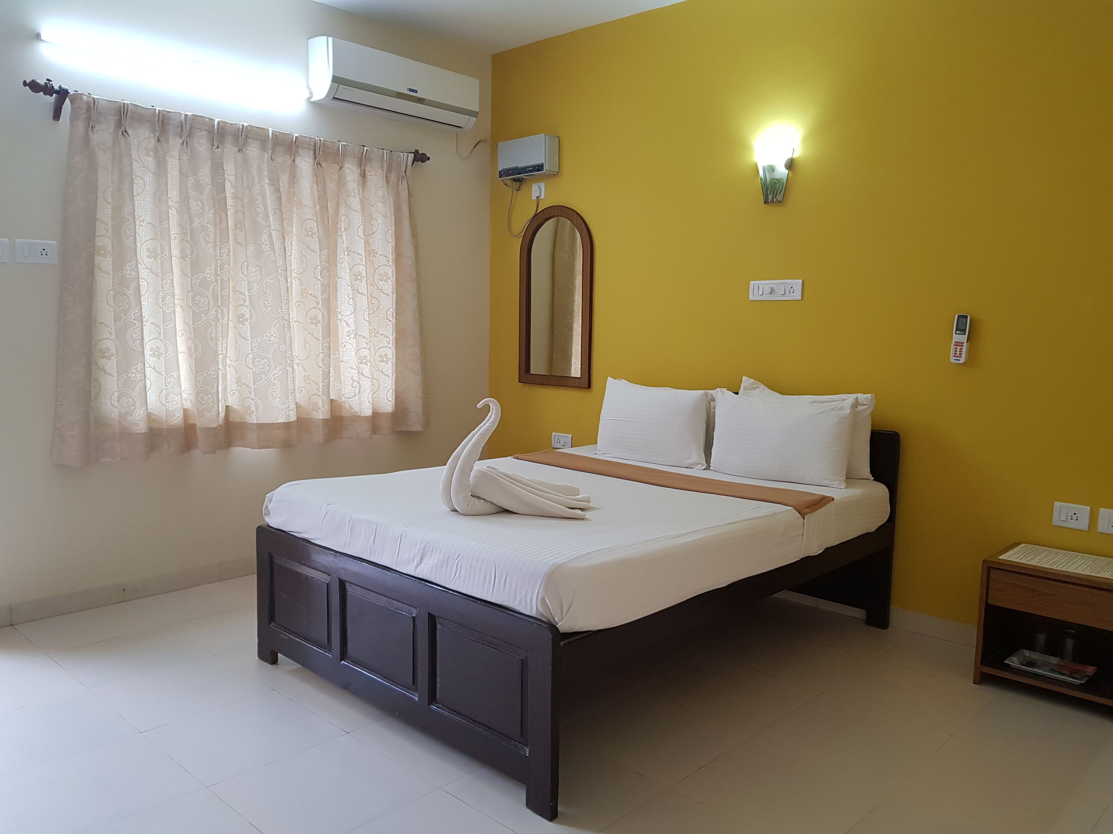 Habitación de hotel con cama, mesita de noche, unidad de aire acondicionado, luz fijada en la pared y ventana con cortinas.