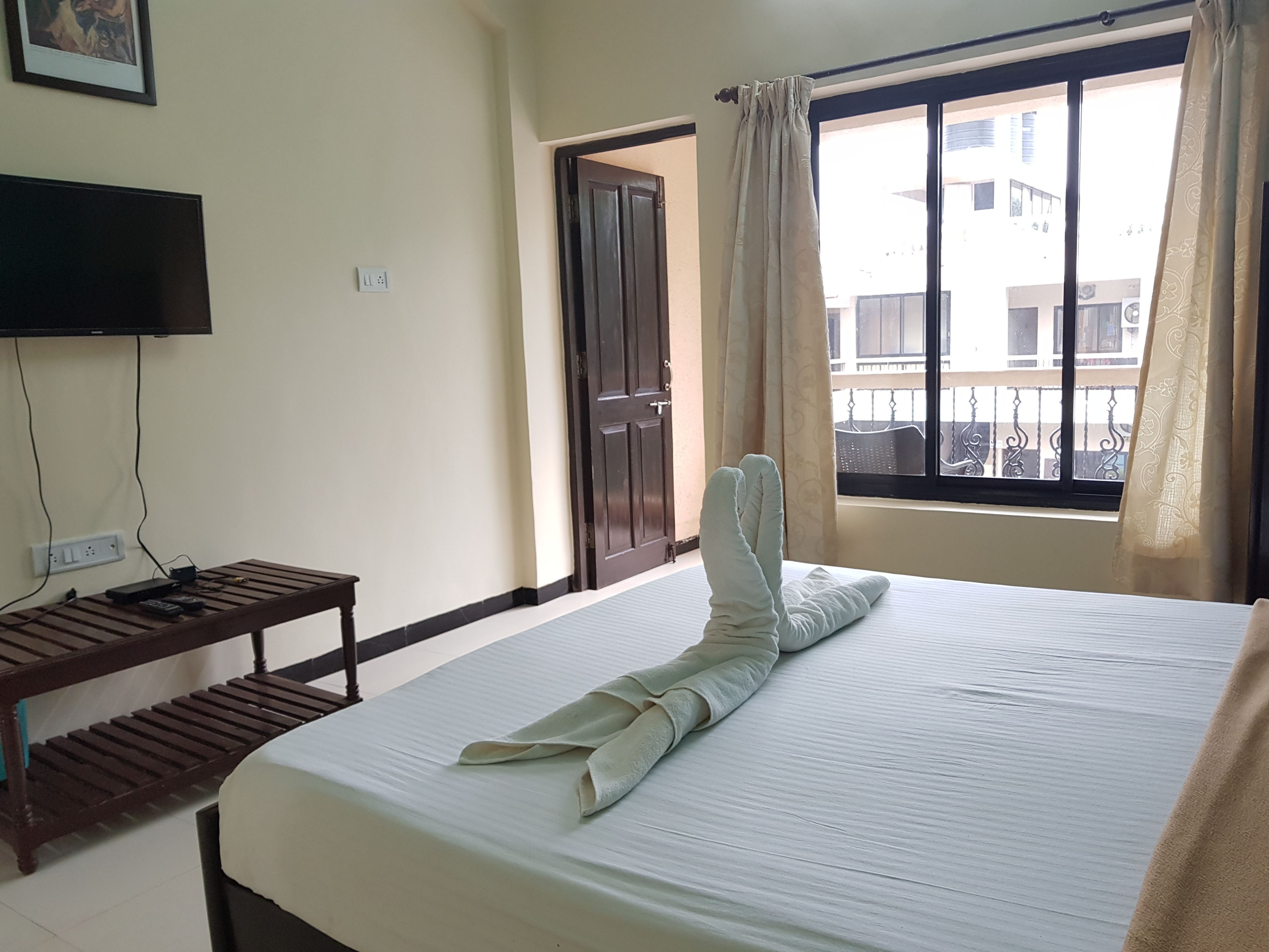 A hotel room with a bed, a TV mounted on the wall, a wooden nightstand, and a window with curtains.