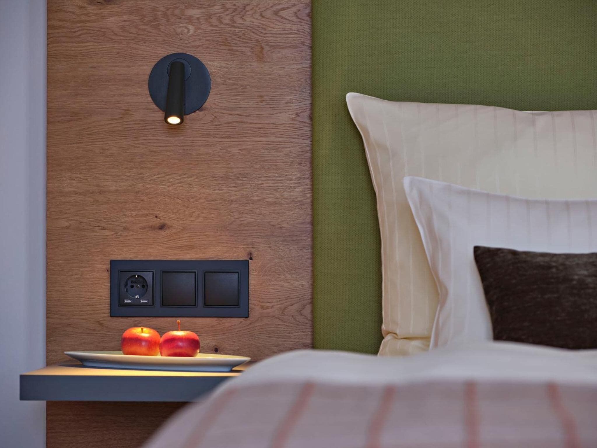 Ein ordentlich bezogenes Bett mit weißen und braunen Kissen, ein Nachttisch mit zwei Äpfeln und eine Wandleuchte.