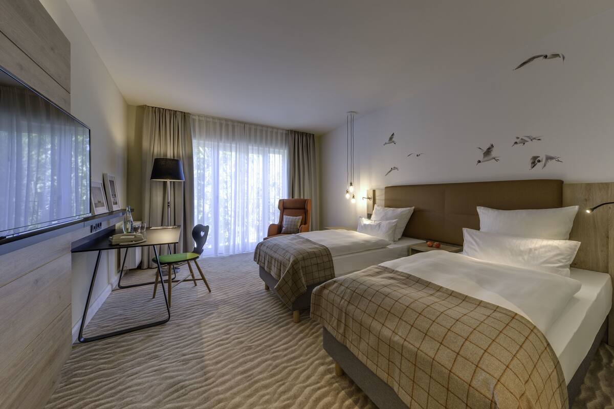 Ein Hotelzimmer mit zwei Betten, einem Schreibtisch mit Stuhl, einer Lampe und einem Fenster mit Vorhängen.