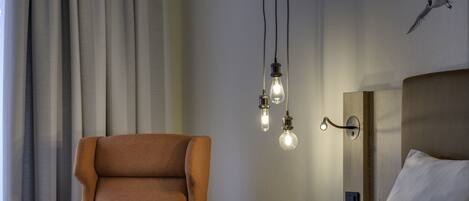 A modern hotel room with a large bed, a plush armchair, and a wall-mounted light fixture.