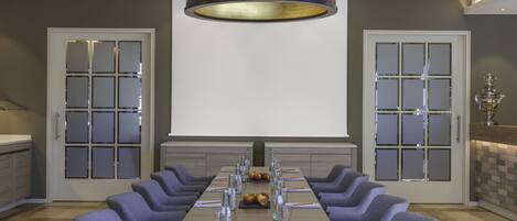 A conference room with a long table, chairs, a projector screen, and a decorative vase.
