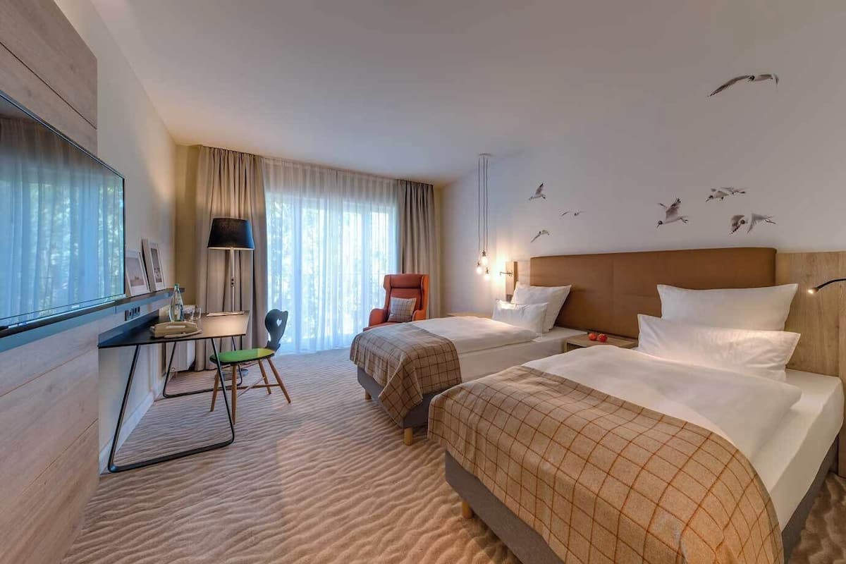 Ein Hotelzimmer mit zwei Betten, einem Schreibtisch, einem Stuhl und einem großen Fenster mit Vorhängen.