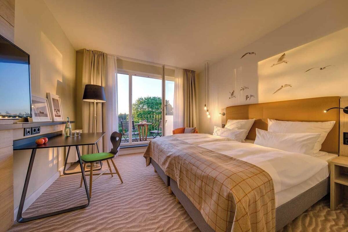 Ein Hotelzimmer mit Bett, Schreibtisch mit Stuhl, einem Balkon mit Tisch und Stühlen sowie Blick ins Freie.