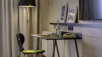 A hotel room with a desk, a chair, a lamp, a telephone, and framed pictures on the wall.