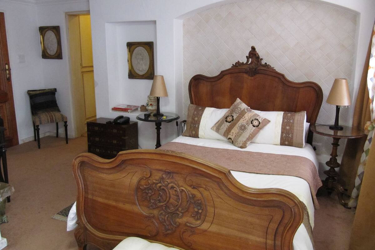 A bedroom with a wooden bed, two bedside tables with lamps, a chair, and two framed pictures on the wall.