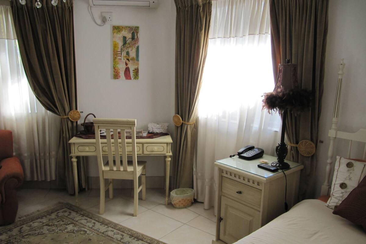 A hotel room with a bed, a desk with a chair, a small table with a phone, a lamp, and a painting on the wall.