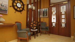 A room with a wooden door, two chairs, a small table with a vase of flowers, a ship's wheel on the wall, and framed pictures on the wall. - Hotel Fabius (Bellaria-Igea Marina)