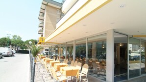 An outdoor seating area with tables and chairs, a glass entrance, and a building with a yellow sign. - Hotel Fabius (Bellaria-Igea Marina)