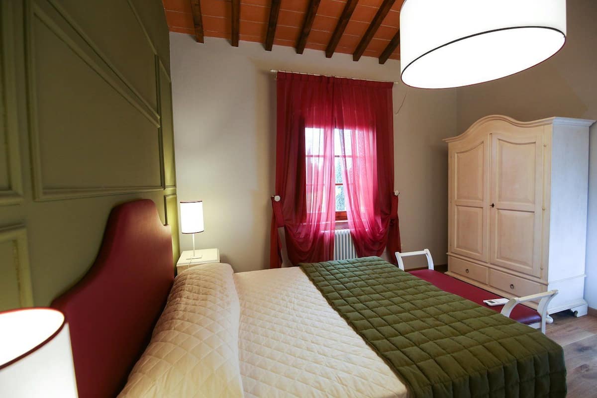 Un dormitorio con cama, mesita de noche, lámpara, armario y ventana con cortinas rojas.