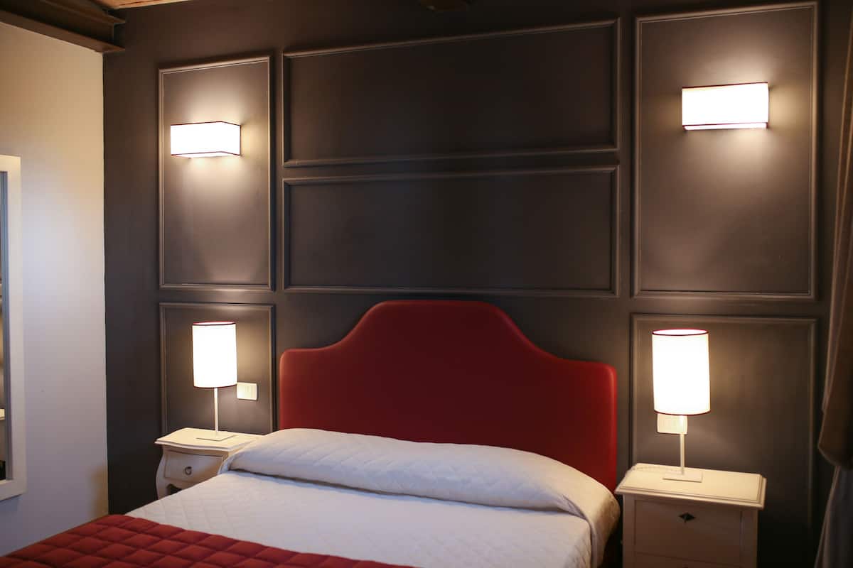 Una habitación de hotel con un cabecero tapizado en rojo, una colcha blanca y dos lámparas de noche.