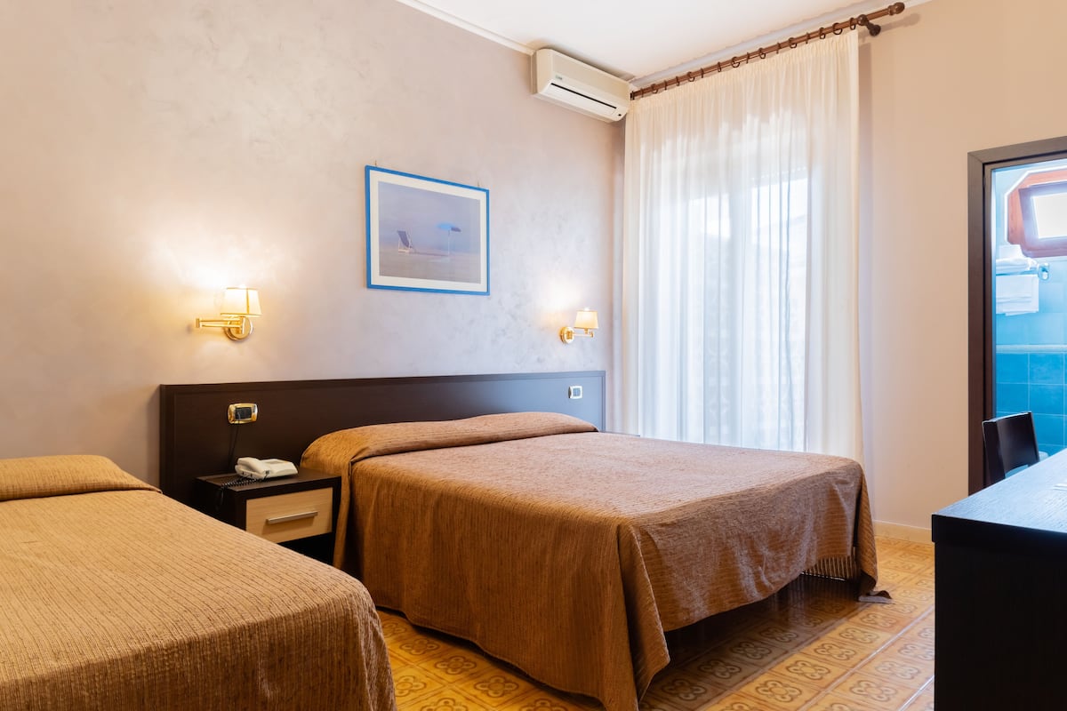 A hotel room with two single beds, a nightstand with a phone, wall-mounted lamps, a framed picture, and a window with curtains.