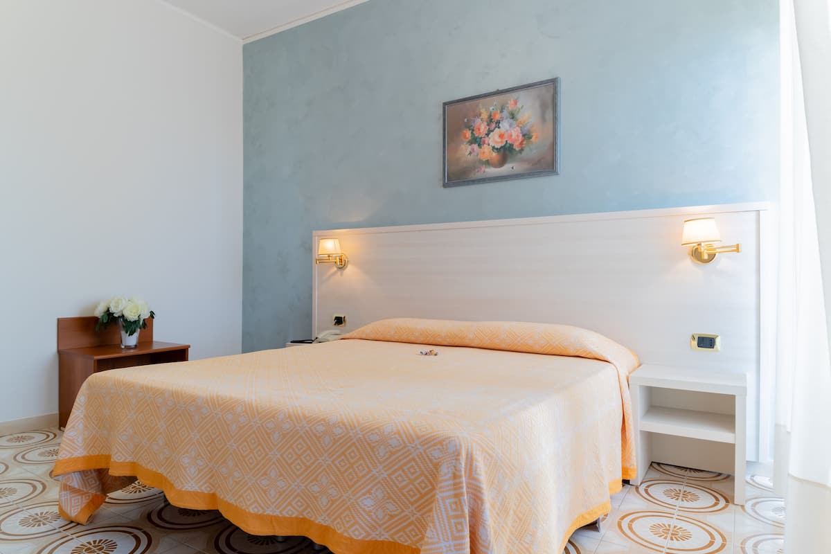 A hotel room with a bed, bedside lamps, a nightstand with a vase of flowers, and a painting on the wall.