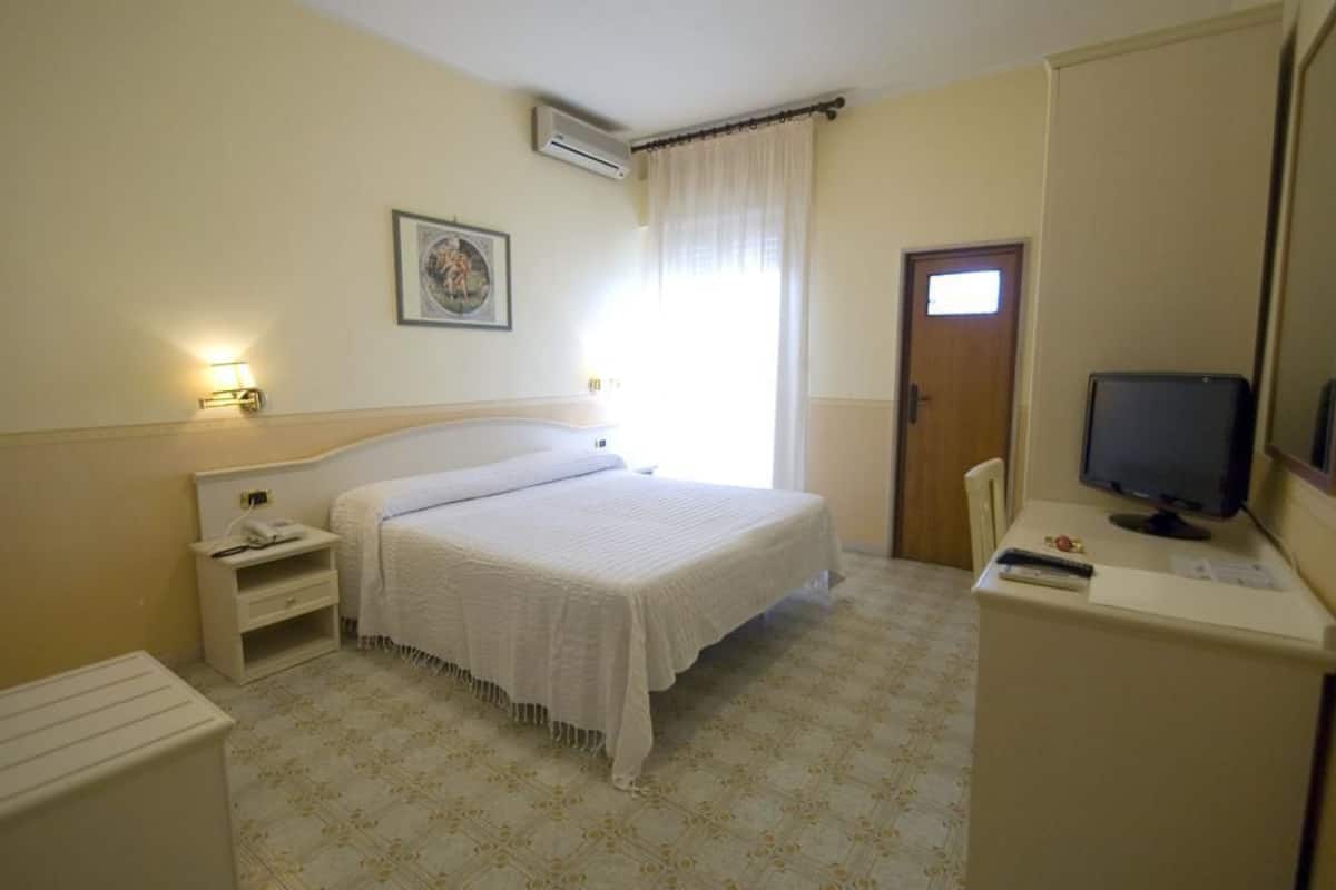 A hotel room with a bed, bedside table, lamp, TV, and a door.