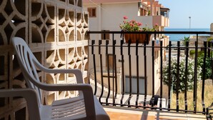 Un balcon avec une chaise en plastique blanche, un pot de fleurs orné de fleurs roses et une vue sur la mer et les bâtiments voisins.