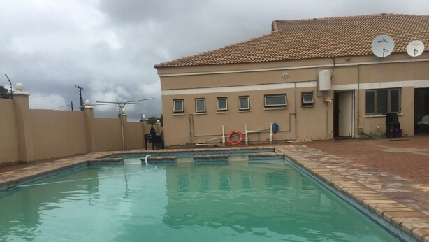 A swimming pool with a tiled deck, a lifebuoy, and a building with satellite dishes in the background. - Warm Hands Hotel (Kanye)