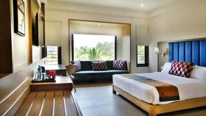 A hotel room with a bed, a sofa, a desk with a tray of items, and a window with a view of greenery. - Purple Cloud Hotel (Bengaluru)