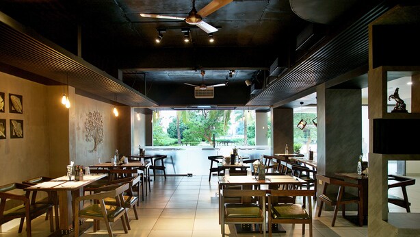 A restaurant with a modern interior, featuring wooden tables and chairs, pendant lighting, and large windows offering a view of greenery outside. - Purple Cloud Hotel (Bengaluru)