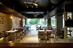 A restaurant with a modern interior, featuring wooden tables and chairs, pendant lighting, and large windows offering a view of greenery outside.