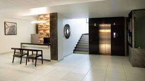 A reception area with a reception desk, two wooden chairs, a wall clock, and a staircase leading to an elevator. - Purple Cloud Hotel (Bengaluru)