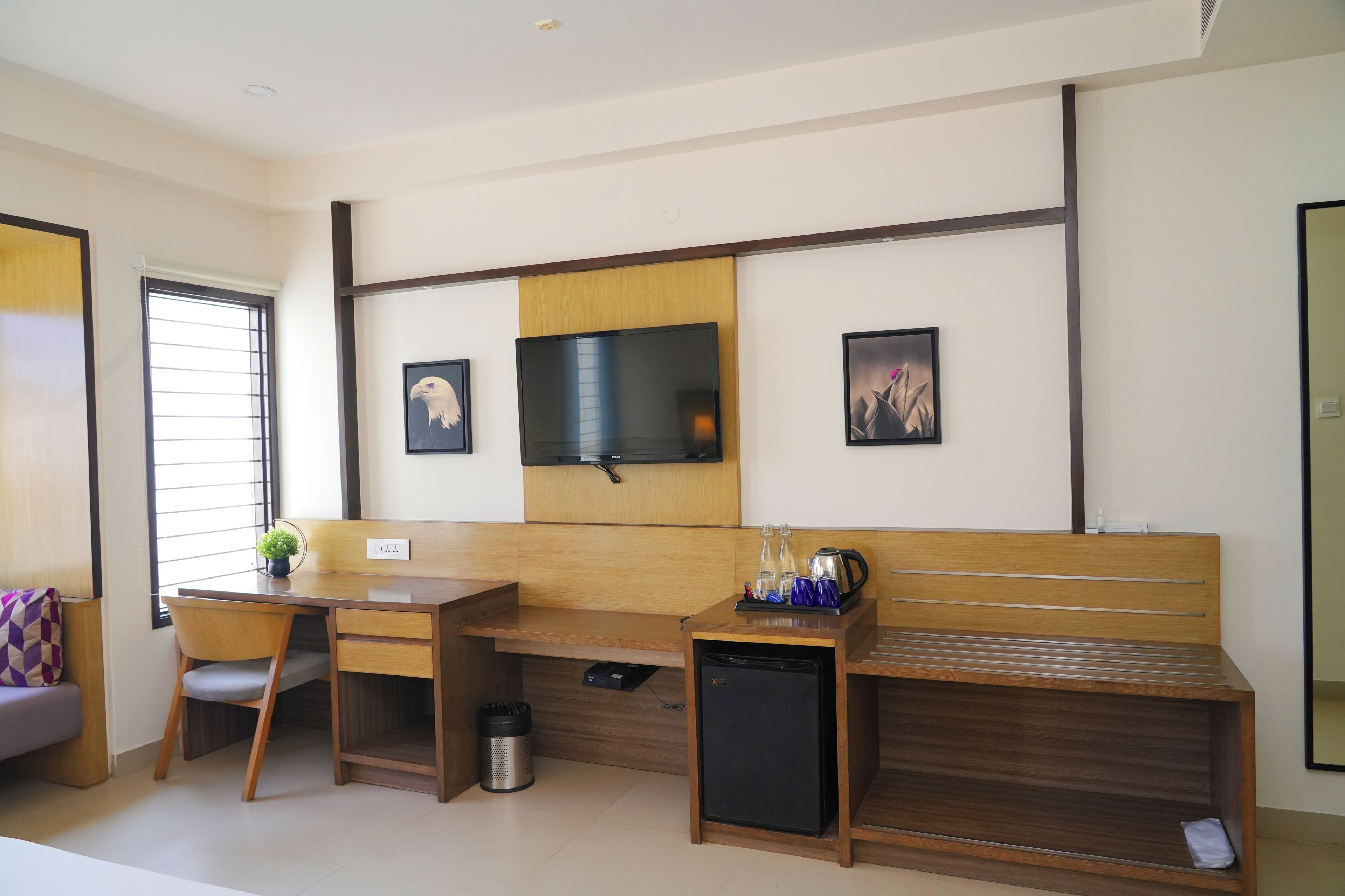 A modern hotel room with a wooden desk, a small refrigerator, a bench, a TV mounted on the wall, and a window with blinds.
