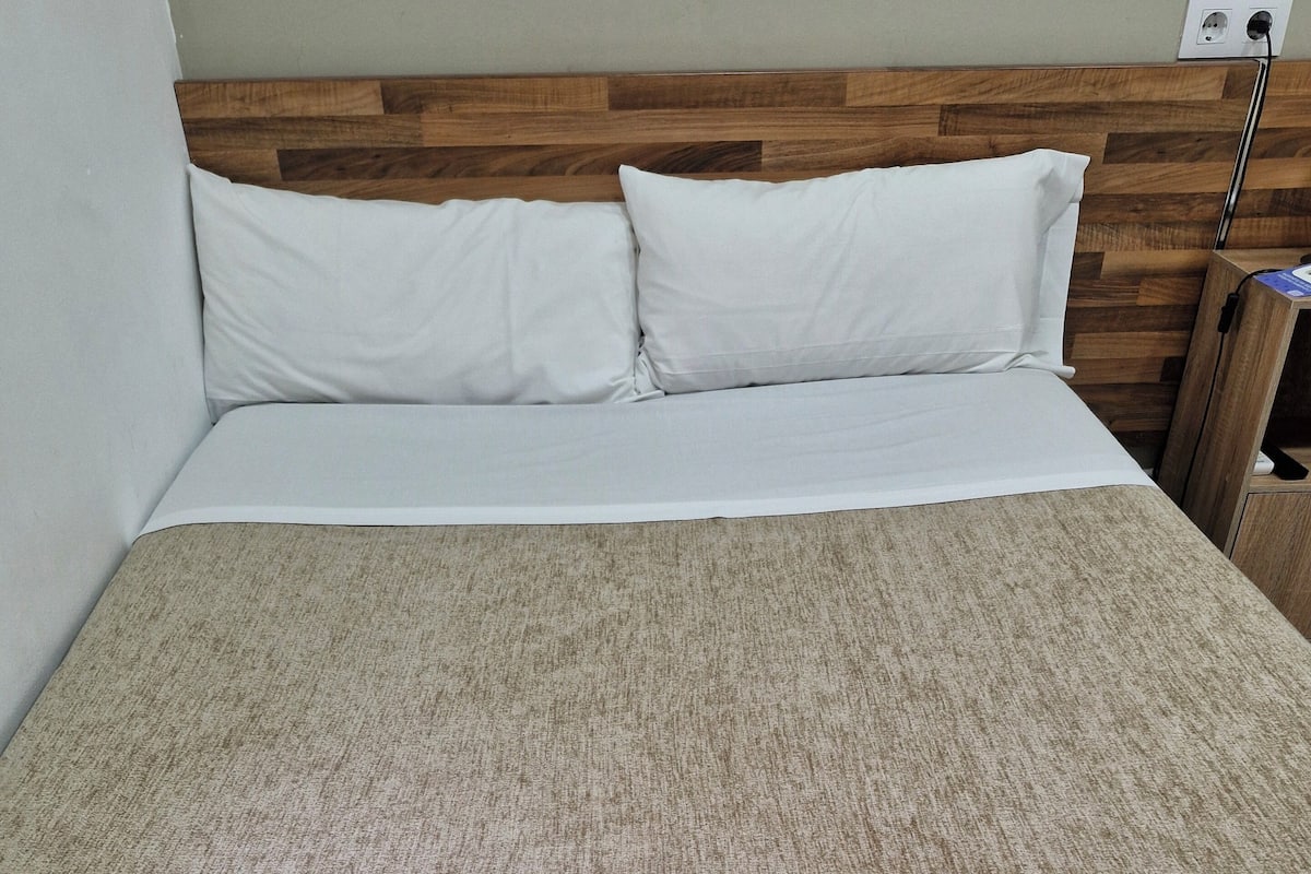 A neatly made bed with white linens, a wooden headboard, and a beige textured bedspread. Two folded towels with toiletries are placed on the bed.