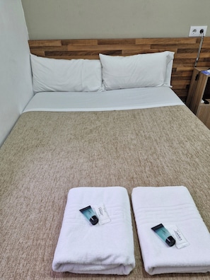Cama bem arrumada com lençóis brancos, cabeceira de madeira e colcha texturizada bege. Duas toalhas dobradas com artigos de higiene pessoal estão sobre a cama.