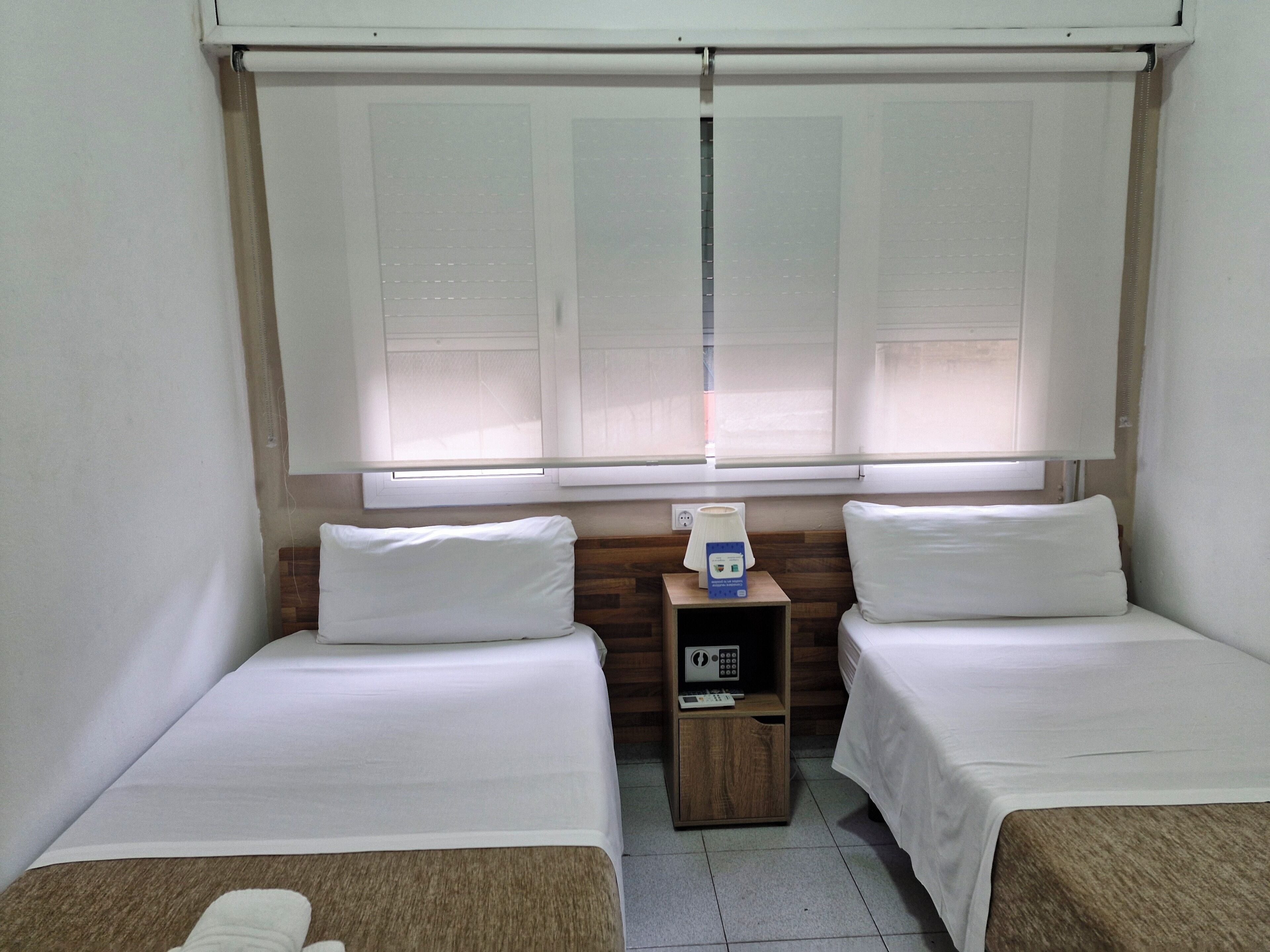 Habitación de hotel con dos camas individuales, una mesita de noche de madera con radio y una ventana con persianas.