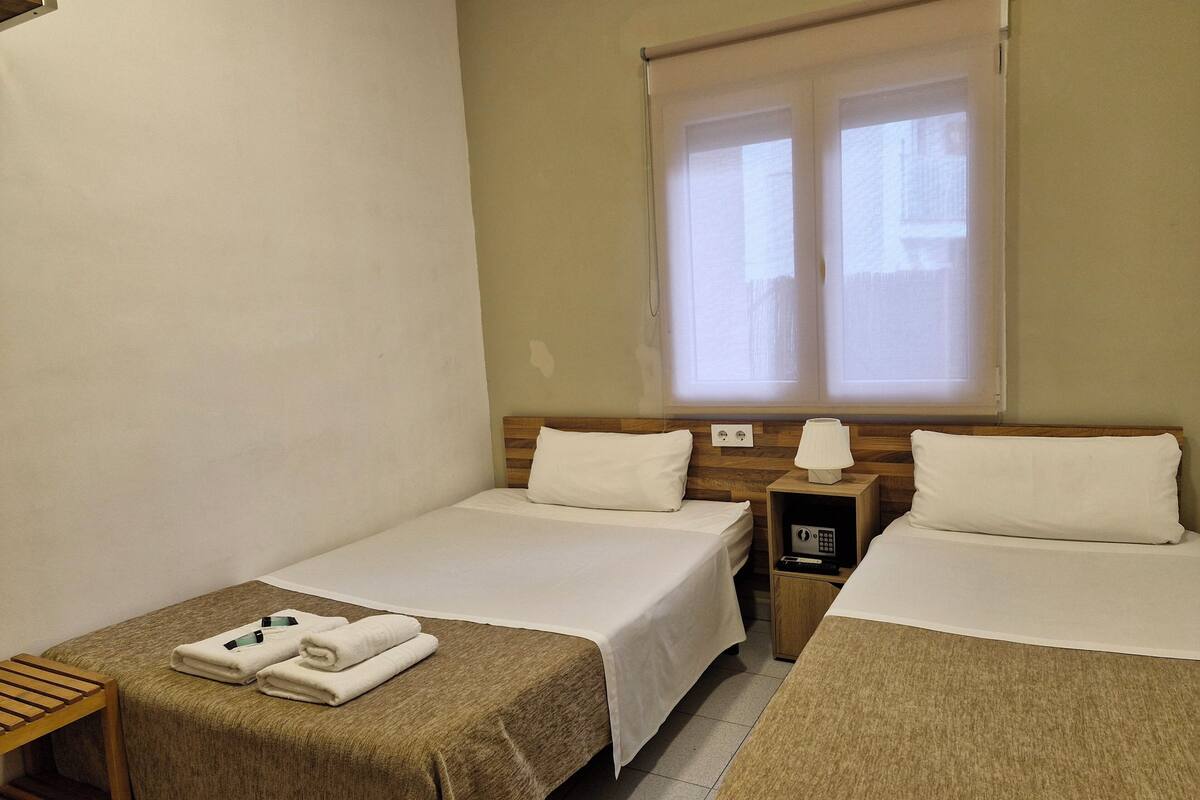 A hotel room with two single beds, a wooden nightstand with a lamp, a small table with towels, and a window with blinds.