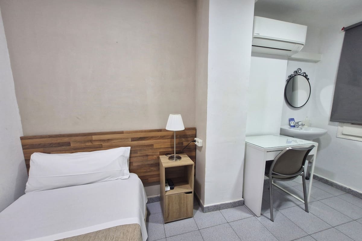 A hotel room with a bed, a desk, a chair, a lamp, a small shelf, a mirror, a sink, and an air conditioning unit.