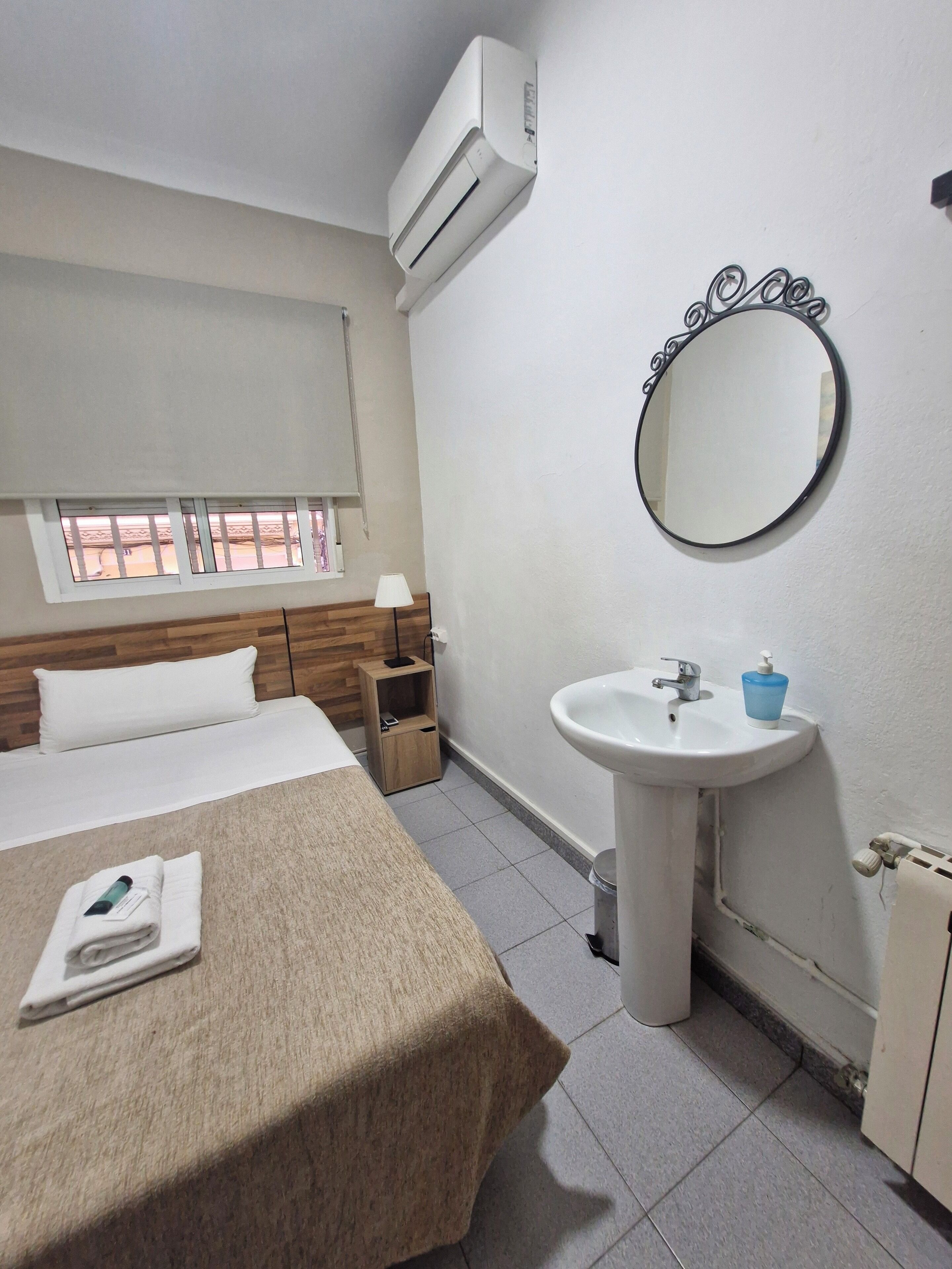 A hotel room with a bed, a bedside table with a lamp, a round mirror, a sink, and an air conditioning unit.