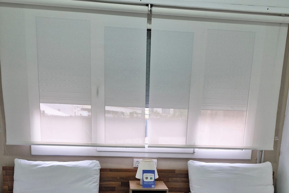 A room with two beds, a nightstand with a lamp, and a window with blinds.