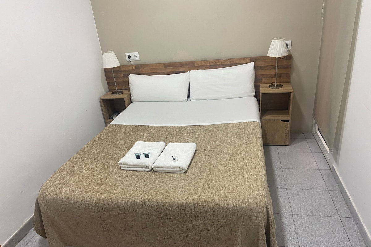 A neatly made bed with a beige bedspread and two folded towels, a wooden headboard, two bedside lamps, and a small wooden nightstand with a lamp.