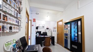 A reception area with a laptop on a desk, a bulletin board with various notices, a vending machine, and a door with a sign. - Hostal Elkano (Barcelona)