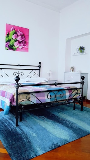 A bedroom with a bed, a blue rug, a white dresser, a nightstand, a lamp, and a pink flower painting.