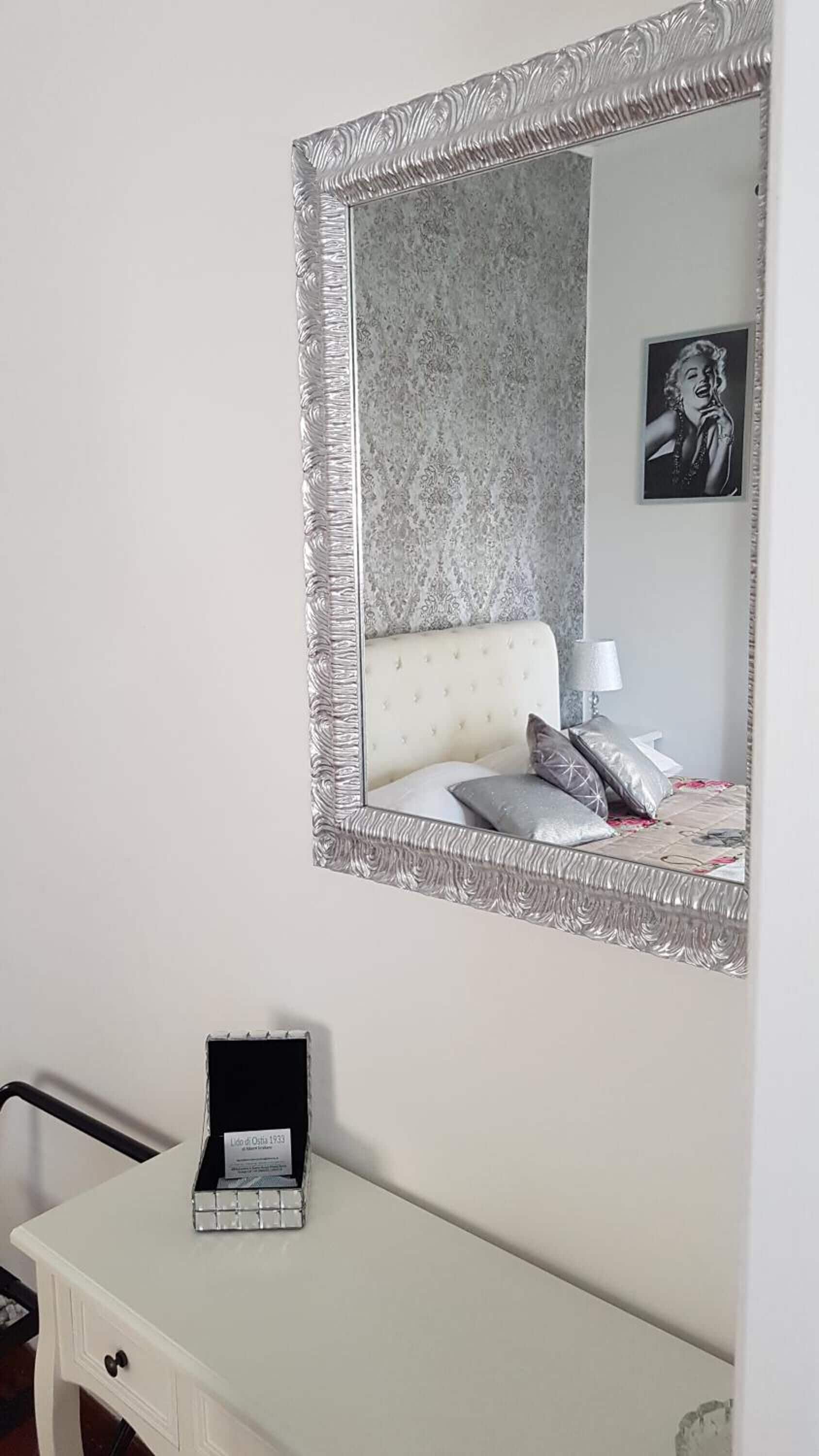 A bedroom with a large ornate mirror, a bed with a floral bedspread, a bedside table with a lamp, and a dresser with a jewelry box.