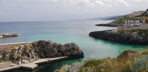 Vista costera con muelle rocoso, aguas turquesas cristalinas y un edificio distante en una colina.