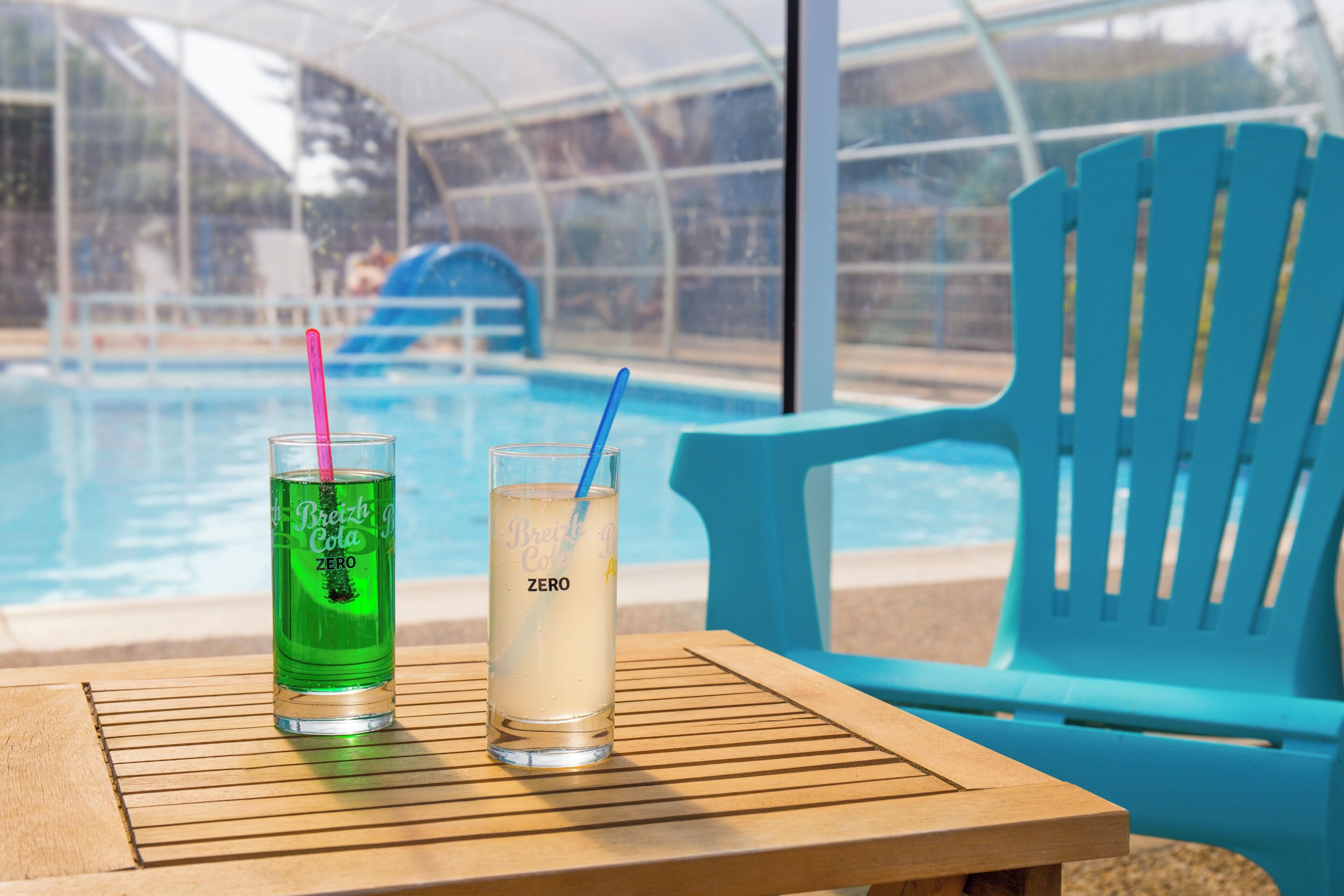 Dos vasos de bebidas sobre una mesa junto a una piscina, con una silla azul y una estructura de techo transparente al fondo.