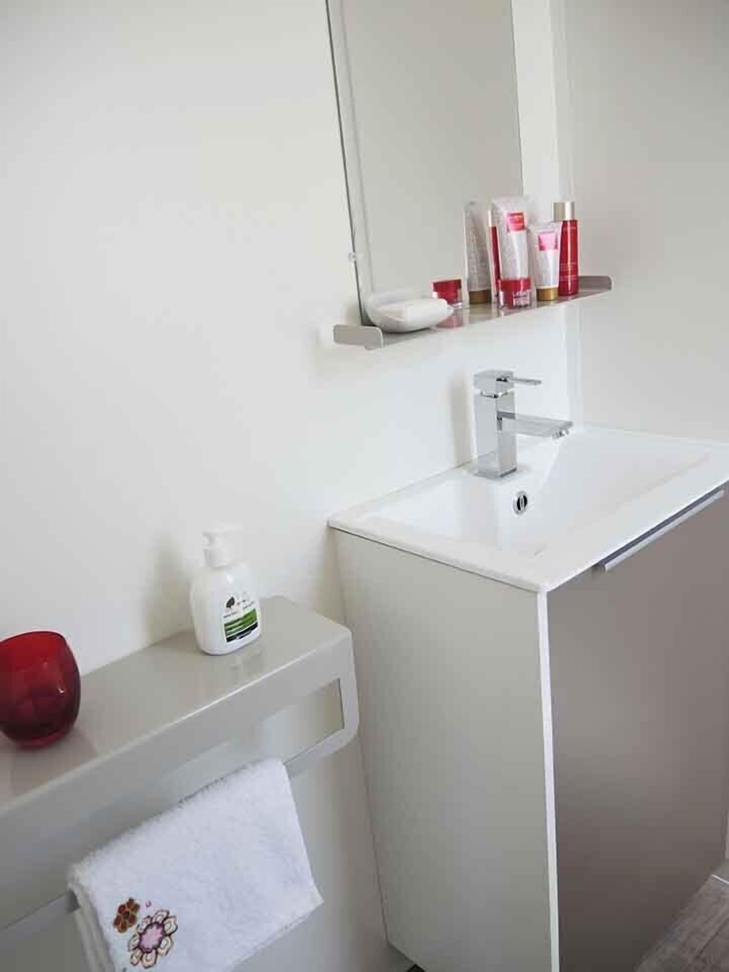 Un baño con un lavamanos blanco, un espejo, un perchero para toallas y un estante con artículos de tocador.