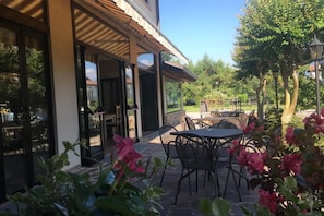 Een terras met tafels en stoelen, een overdekt zitgedeelte en uitzicht op de omliggende begroeiing.