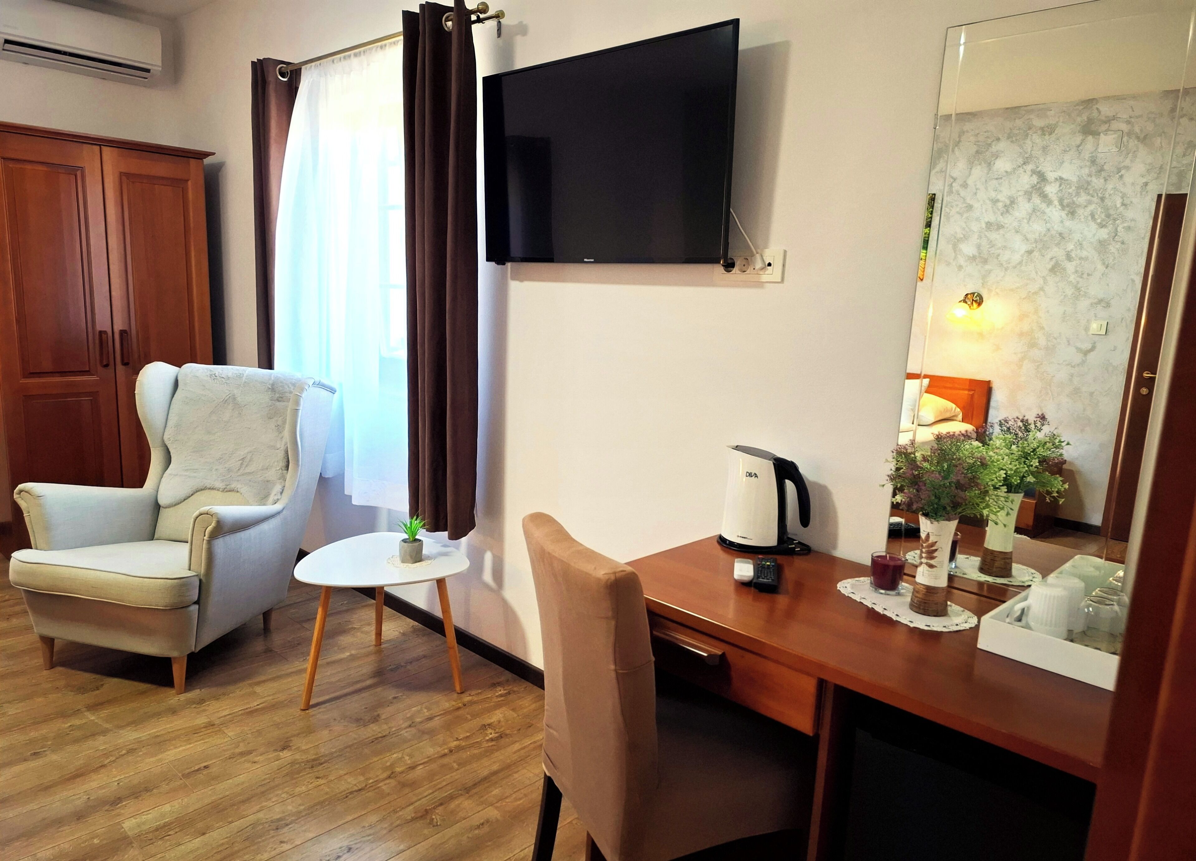 Ett hotellrum med en garderob i trä, en beige fåtölj, ett litet runt bord med en växt, en kaffebryggare och en tv som är monterad på väggen.