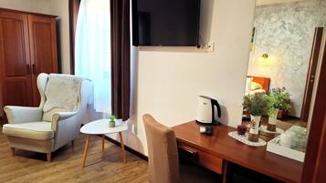 Ett hotellrum med en garderob i trä, en beige fåtölj, ett litet runt bord med en växt, en kaffebryggare och en tv som är monterad på väggen.