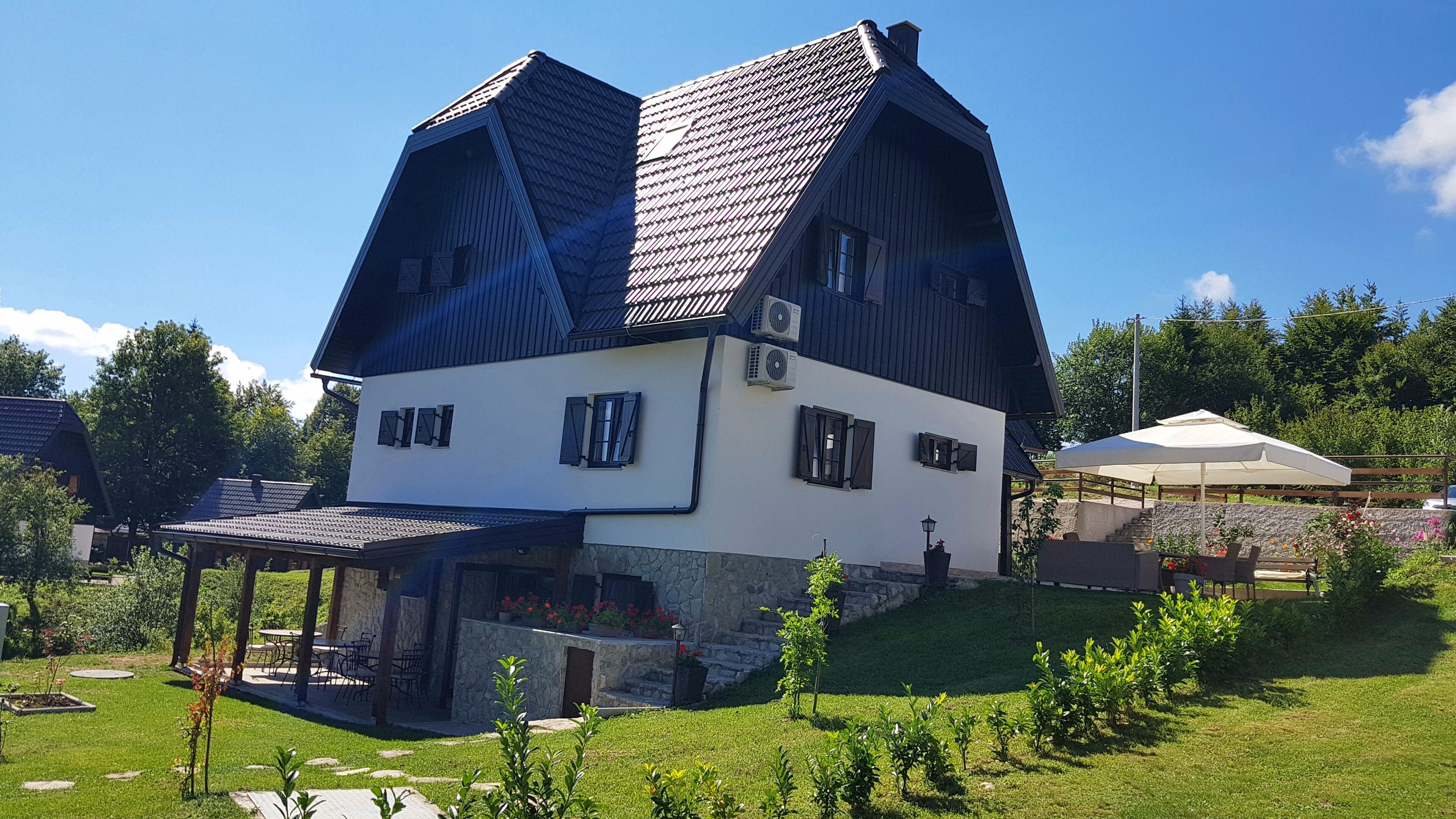 Uma casa de dois andares com telhado inclinado, um alpendre com área de estar ao ar livre e uma parede de pedra.