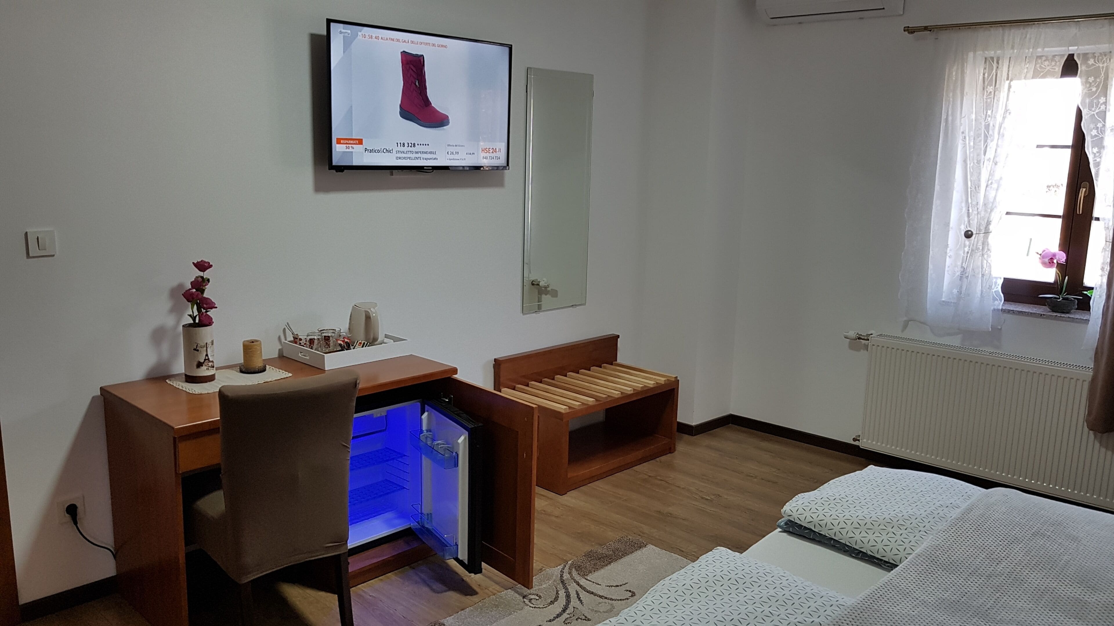 Een hotelkamer met een houten bureau, een stoel, een bed met wit beddengoed, een klein houten bankje, een aan de muur bevestigde televisie en een raam met uitzicht.