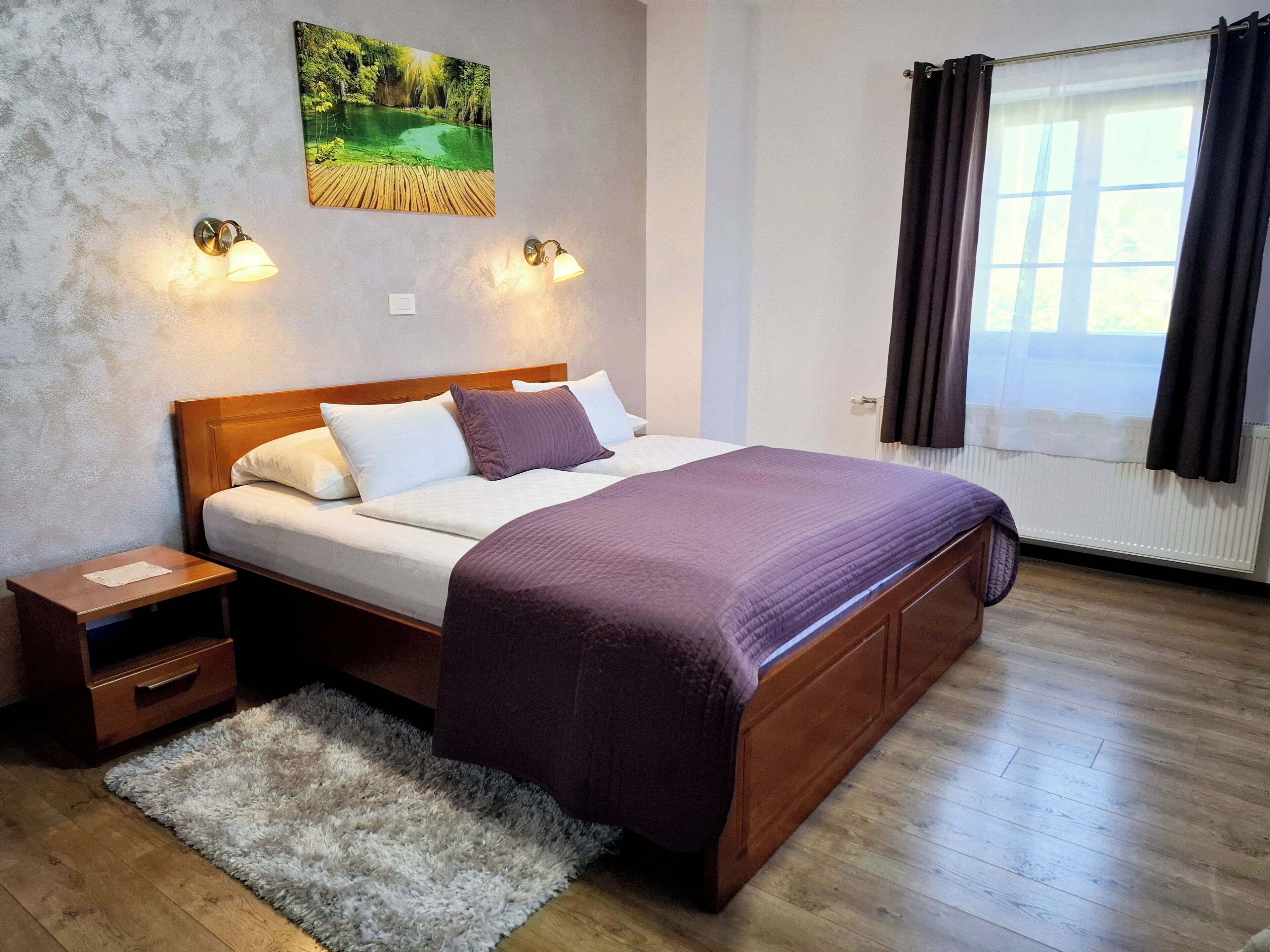 Una cama con ropa de cama bien ordenada, un cabecero de madera, dos mesitas de noche, un tapete mullido y un cuadro de un paisaje en la pared.