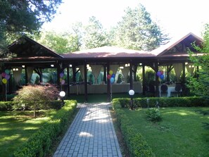 A covered outdoor area with tables and chairs, surrounded by greenery and trees. - Hotel Baneasa Parc (Bucharest)